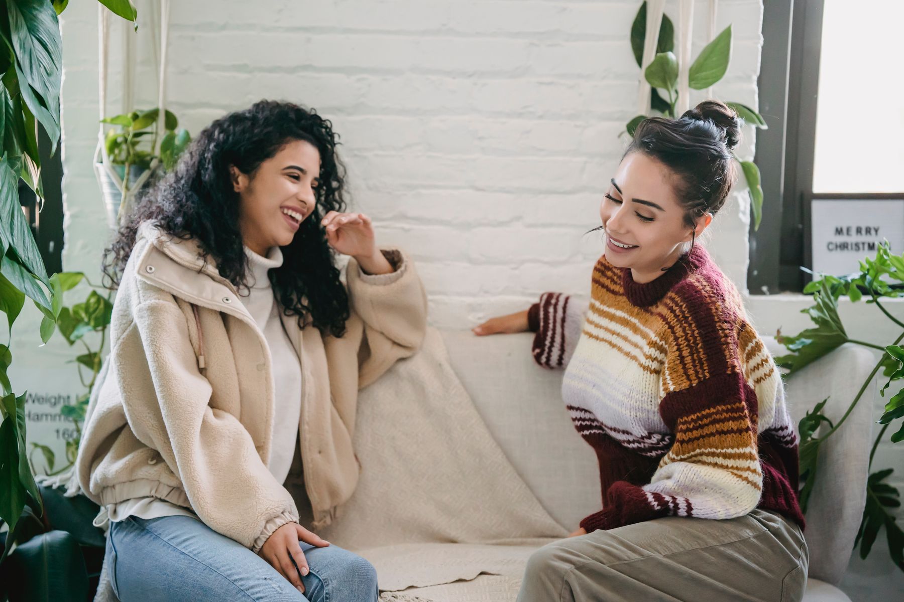 Two women sharing a relaxed conversation on a sofa in a bright, plant-filled room